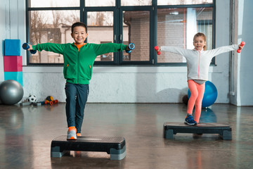 Multicultural children with outstretched hands holding dumbbells and doing exercise on step platforms in gym