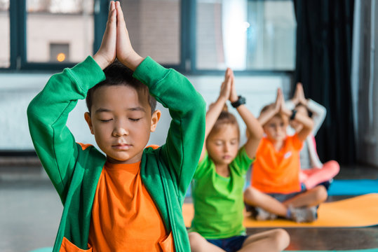 Selective focus of multicultural children meditating with clenched hands over heads in gym