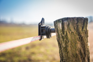 Black electric fence insulator on a fence post. Pole made of tree branch with bark. Meadow and blue...