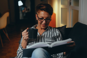 woman relaxed on sofa reading book and drinking coffee or tea at her home, evening scene