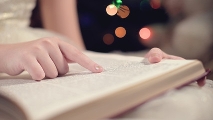 Close-up soft focus the hand of a little girl leads along the lines of a book while reading a New Year's fairy tale amid the lights of a Christmas tree. The concept of a fabulous mood