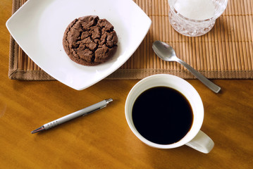 Black coffee in white cup in messy arrangement on table, with chocolate cookie on white plate, teaspoon, pen, bamboo placemat and sugar-bowl on table, indoors, top view