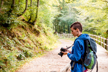 Serious youthful female hiker enjoying vacation and taking picture on professional camera while standing on wooden pathway in forest