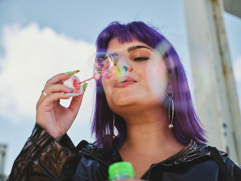 Fashion woman with purple hair smiling and blowing bubbles holding bottle with closed eyes with manicured hands in bright day