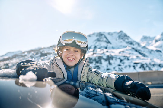 Delighted boy in helmet with goggles smiling and rolling snowball while peeking out vehicle window on sunny day on ski resort