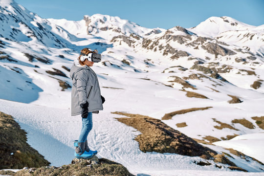Side view of happy funny boy in outwear standing on snow and exploring virtual reality on sunny winter day in mountains