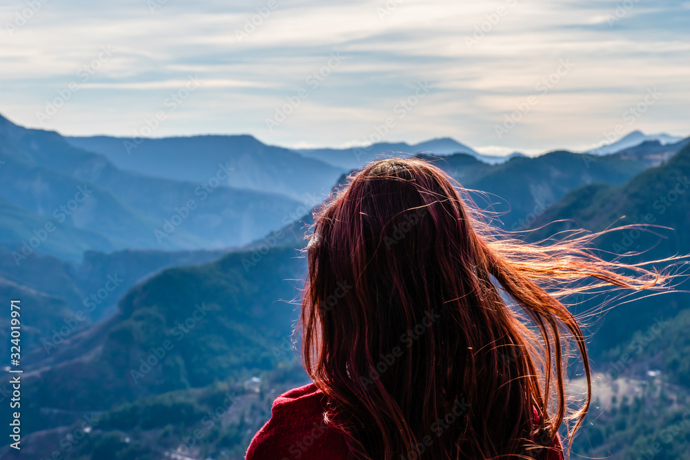 Wall mural a close-up back view of a young redhead woman with her long hair fluttering in front of the mountain