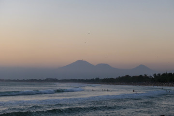 Golden hour on Seminyak Beach, Bali, Indonesia. The sky has pinkish color. The beach bay is washed by gentle waves, There are high volcanic mountains in the back. Idyllic and relaxing moment
