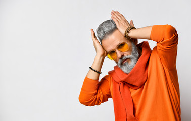 Old-aged male in orange sweater and scarf, sunglasses, bracelets. Touching his head by hands, posing isolated on white. Close up