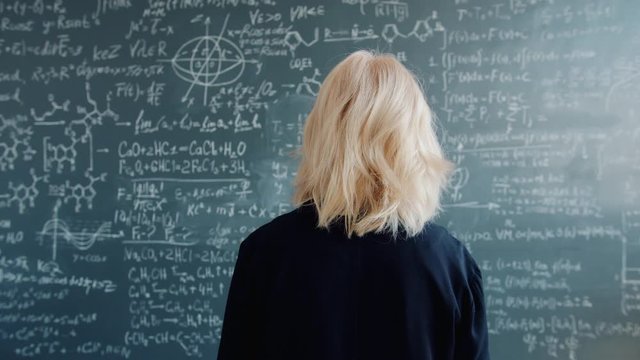Adult blonde is walking to blackboard with scientific formulas in university classroom looking around thinking about science. People and education concept.