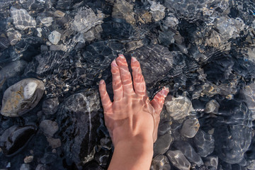 A young woman's hand underwater touching pebbles under clean transparent water of a mountain river with sunlight reflecting and flickering