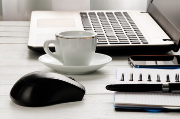 Cup of coffee, laptop with mouse, notebook with pen and smartphone on white desktop