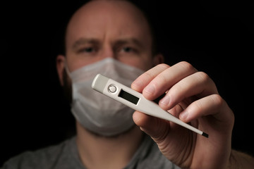 A young man in a medical mask holds a thermometer in his hand on black background. Thermometer close up. Seasonal infection, colds and runny nose, coronavirus. Health and healthcare concept