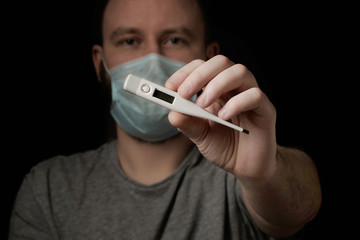 A young man in a medical mask holds a thermometer in his hand on black background. Thermometer close up. Seasonal infection, colds and runny nose, coronavirus. Health and healthcare concept