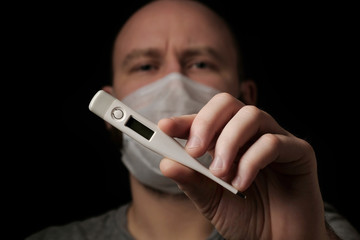 A young man in a medical mask holds a thermometer in his hand on black background. Thermometer close up. Seasonal infection, colds and runny nose, coronavirus. Health and healthcare concept