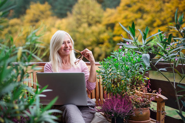 Senior woman with laptop sitting outdoors on terrace, working.