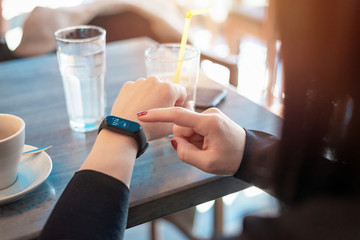 Girl touch smart band, bracelet screen on her arm. Displayed ime and number of steps. Caffee shop in background