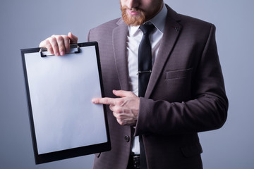 Studio portrait of a young bearded handsome guy of twenty-five years old, in an official suit, holding a folder with a sheet for text. Free space on a gray background. Office Style. Fashion and office