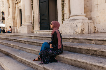 Portrait of beautiful woman with hijab sitting on stairs in front of old church, historical building,  in Dubrovnik, Croatia. She is holding hand fan. 