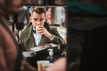 Fashionable young male customer sitting outdoors next to coffee making barista in a mobile coffee shop in a city emporium, wearing green wool coat, white shirt and bow tie, taking a sip of coffee of