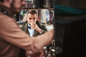 Stylish and elegant young male customer sitting outdoors next to coffee making barista in a mobile coffee shop in a city emporium, wearing green wool coat, white shirt and bow tie, taking a sip of