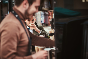 Fashionable young male customer sitting outdoors next to coffee making barista in a mobile coffee shop in a city emporium, wearing green wool coat, white shirt and bow tie, taking a sip of coffee of