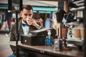 Stylish and elegant young male customer sitting outdoors next to coffee making gear in a mobile coffee shop in a city emporium, wearing green wool coat, white shirt and bow tie, taking a sip of coffee