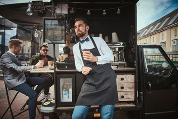 Hipster looking man barista working in a mobile coffee shop outdoors in the city emporium wearing apron and white shirt with bow tie, standing near coffee machine and two customers holding a paper cup