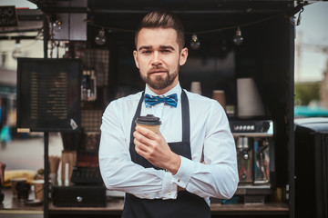 Handsome looking man barista working in a mobile coffee shop outdoors in the city emporium wearing apron and white shirt with bow tie, standing near coffee machine with arms crossed holding a paper