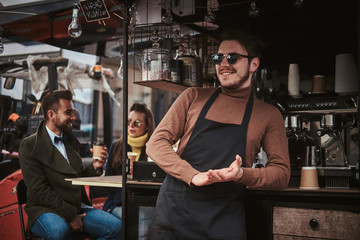 Smiling and elegant male barista standing relaxed outdoors near mobile coffee shop and two elegant looking, chatting customers wearing dark apron, sunglasses and warm wool brown sweater in a