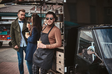 Fototapeta na wymiar Charming and calm male barista standing relaxed outdoors near mobile coffee shop and two elegant looking, chatting customers wearing dark apron, sunglasses and warm wool brown sweater in a combination