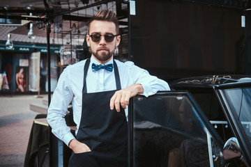 Handsome looking man barista standing near mobile coffee shop outdoors in the city emporium wearing apron, white shirt with bow tie and sunglasses, looking confident and calm