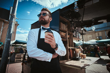 Handsome looking man barista working in a mobile coffee shop outdoors in the city emporium wearing apron and white shirt with bow tie, standing near coffee machine with arms crossed holding a paper - obrazy, fototapety, plakaty