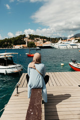 woman with hijab sitting on deck by the sea.  Female tourist exploring Cavtat. She is sitting turned back and looking in the old city. 