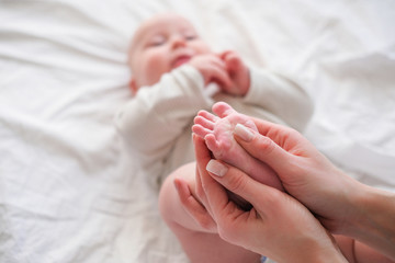 Baby feet in mother hands. Young caucasian woman makes massage for happy infant baby on white bed at home. Babycare, sport and happy motherhood