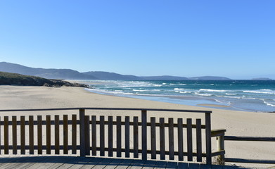 Beach with wooden boardwalk and handrail. Wild sea with waves and blue sky. Arteixo, Coruña, Galicia, Spain.