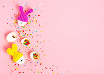 Easter greeting card. Decorative eggs with painted cheerful faces and crocheted ears in the form of bunnies, colorful confetti in eggshell on the pink background. Copy space, top view