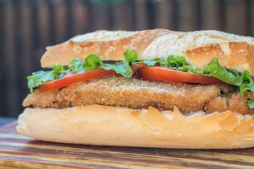 Tasty big fried chicken sandwich on french bread with tomatoes and lettuce on wooden table and rustic background. Top view.