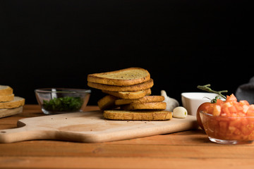 Grilled slices of bread lying on a wooden board surrounded by ingredients for bruschetta dark background warm light