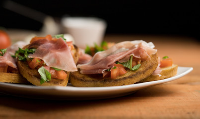 Bruschetta with tomato, basil, garlic and Italian ham on a white plate surrounded by ingredients, warm light closeup