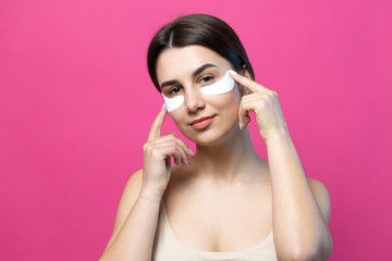 Close up portrait of pretty attractive girl with naked shoulders using patches under eyes. Standing over pink background.