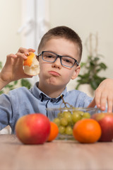 Chłopiec w niebieskiej koszuli i okularach siedzi przy stole i trzyma w dłoni jabłko które je ze smakiem. © Tomasz