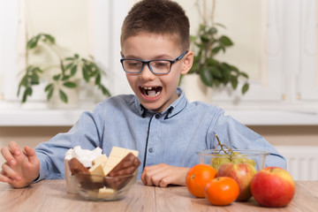 Chłopiec w okularach siedzi przy stole i z zadowoloną miną wpatruje się  w słodycze. Obok leżą owoce mandarynki, jabłka i winogron. - obrazy, fototapety, plakaty