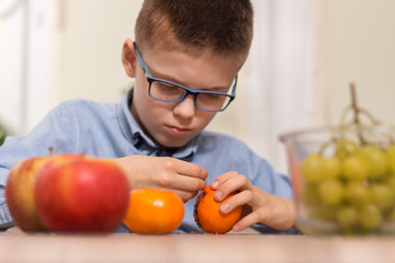 Chłopiec w okularach ze skupieniem wbija w owoc mandarynki goździki. Zdobienie owoców mandarynki goździkami przez chłopca w wieku szkolnym. Na pierwszym planie na blacie leżą owoce jabłka i mandarynki