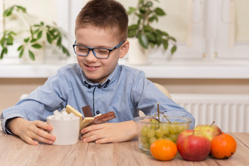 Chłopiec w wieku szkolnym z uśmiechem wpatruje się i trzyma naczynie ze słodyczami oraz cukiernicę z kostkami cukru. Obok na blacie stołu stoją owoce mandarynki, winogrona i jabłka. - obrazy, fototapety, plakaty