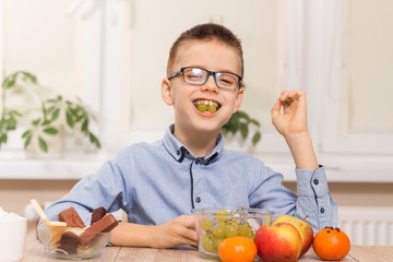Uśmiechnięty chłopiec siedzi przy stole i zjada owoce. Trzyma w zębach owoce winogrona. © Tomasz