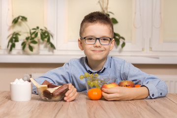 Uśmiechnięty chłopiec w wieku szkolnym siedzi przy stole i przysuwa do siebie owoce. Drugą ręką odsuwa od siebie naczynia ze słodyczami. - obrazy, fototapety, plakaty