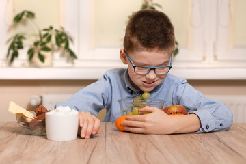 Uśmiechnięty chłopiec w wieku szkolnym siedzi przy stole i przysuwa do siebie owoce. Drugą ręką odsuwa od siebie naczynia ze słodyczami. - obrazy, fototapety, plakaty