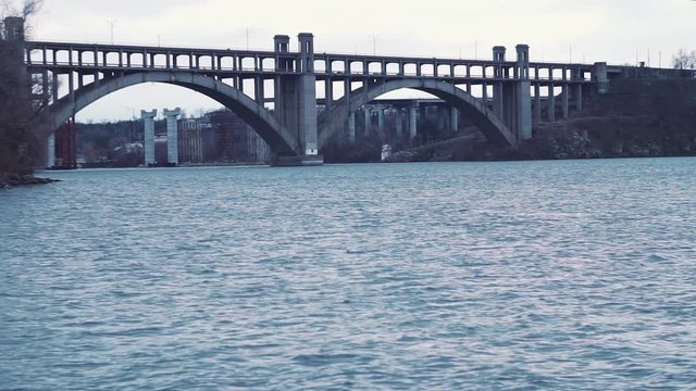 The old arched bridge of the Transfiguration, against the backdrop of the unfinished structures of the new, blue, gray ripples of the Dnieper River, Khortitsa Island. Day cold winter cars drive