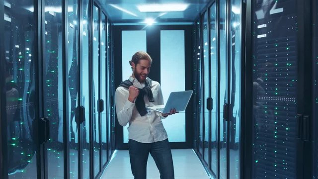 Successful engineer working IT project on laptop smiling happy celebrating good news standing in server room of data center. Technology and people.